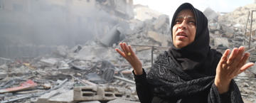 سيدة ترتدي الحجاب تجلس وسط أنقاض المباني المدمرة، ترفع يديها بالدعاء، ويبدو على وجهها الحزن والأسى وسط مشهد الدمار والدخان المتصاعد.