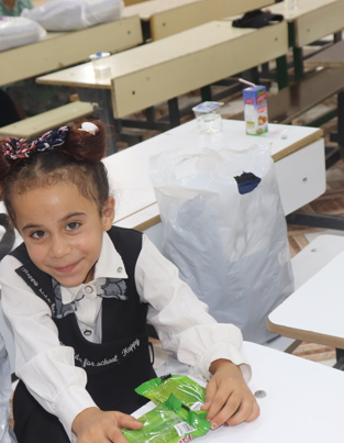 أجر تربية اليتيم وفضل كفالته في الإسلام فتاة صغيرة ذات تسريحة شعر على شكل فيونكة تبتسم للكاميرا أثناء جلوسها على مقعد دراسي في الفصل. ترتدي فستان مدرسي باللونين الأسود والأبيض وتحمل علبة من الوجبات الخفيفة. في الخلفية، يمكن رؤية طلاب آخرين جالسين على مقاعد دراسية مع مستلزمات مدرسية.